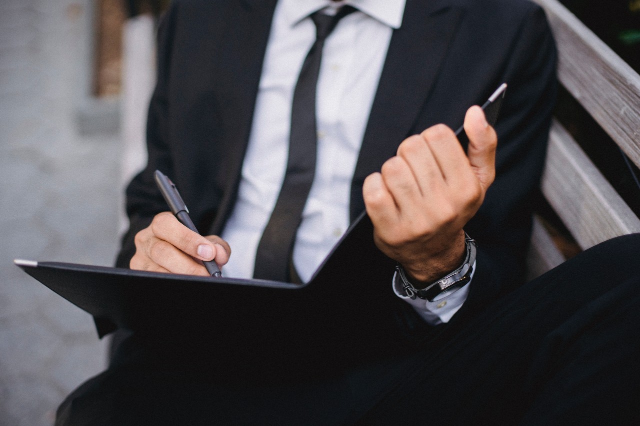 A man in a black suit and tie sits on a bench, holding a folder and pen.