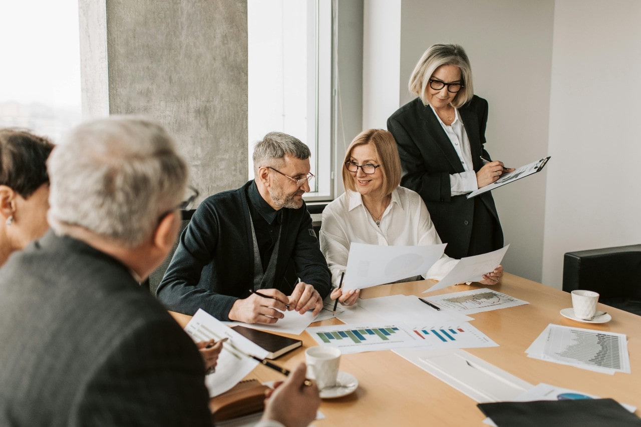 A group of professionals in business attire are gathered around a conference table reviewing charts and documents during a meeting.