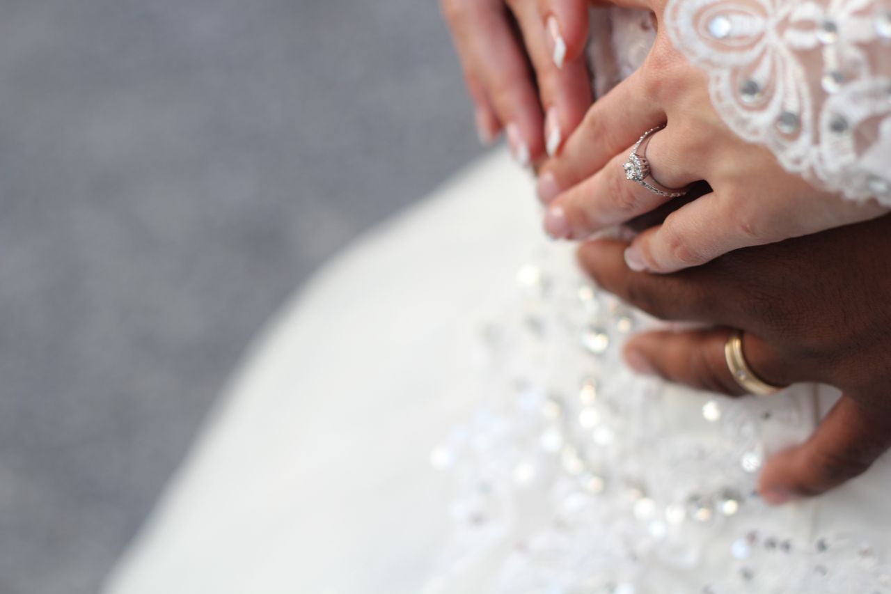 A close-up of a couple's hands intertwined with rings, resting on a white, beaded wedding gown.