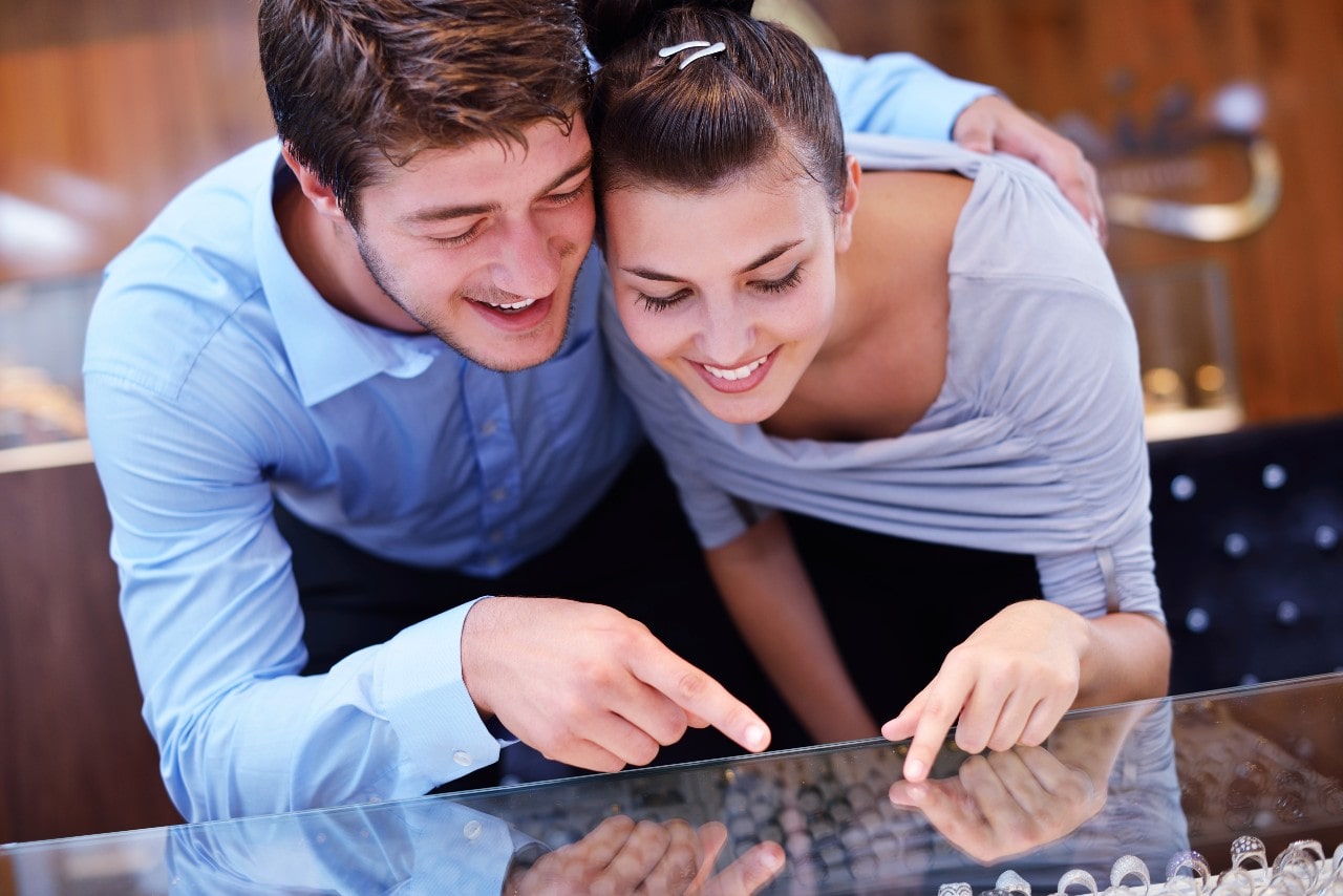 A smiling couple leans over a jewelry store display case, pointing at engagement rings.