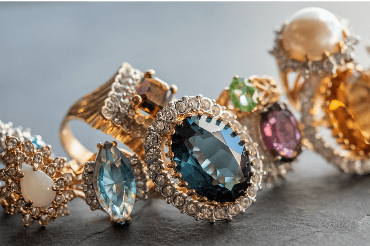 A row of elegant gold rings featuring large, colorful gemstones and sparkling diamonds is displayed against a dark background.