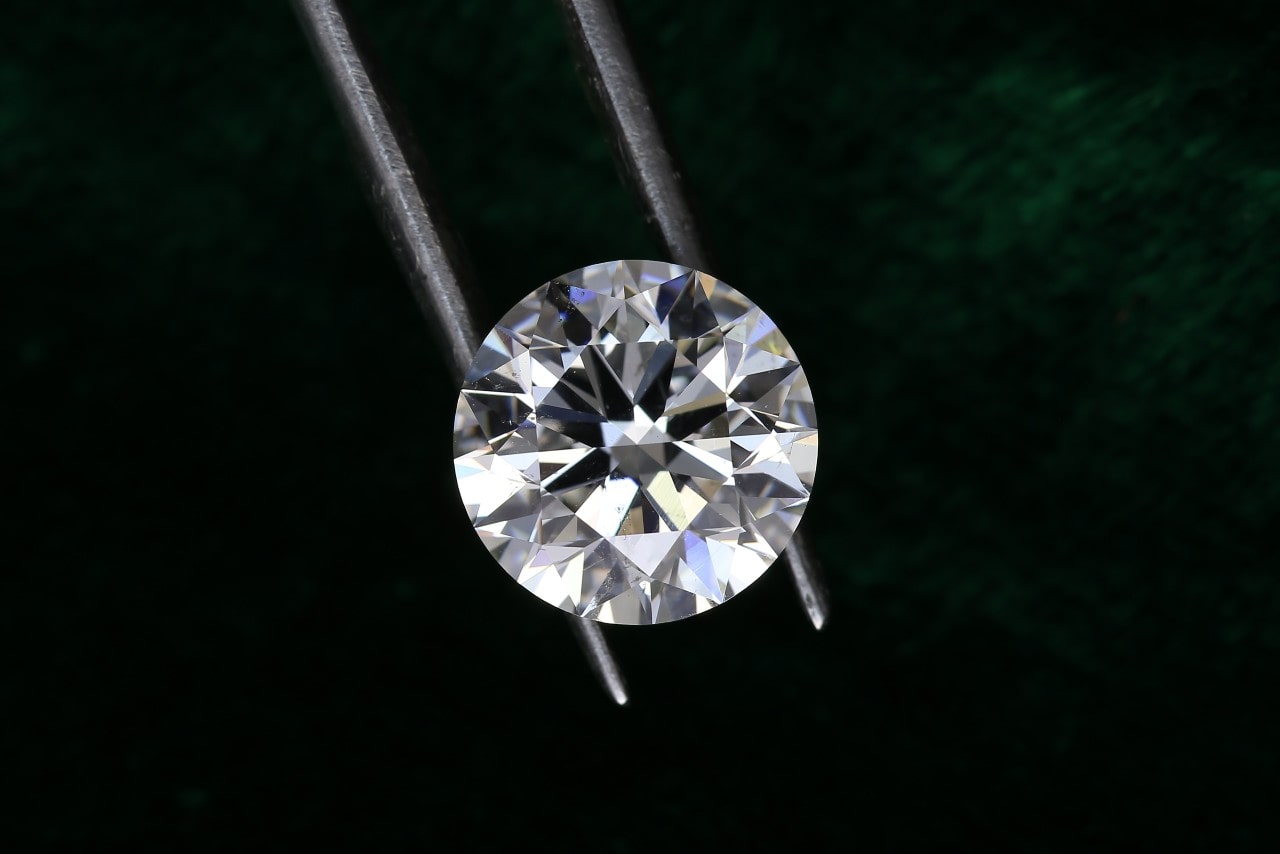 A round-cut diamond is held by metal tweezers against a dark green background