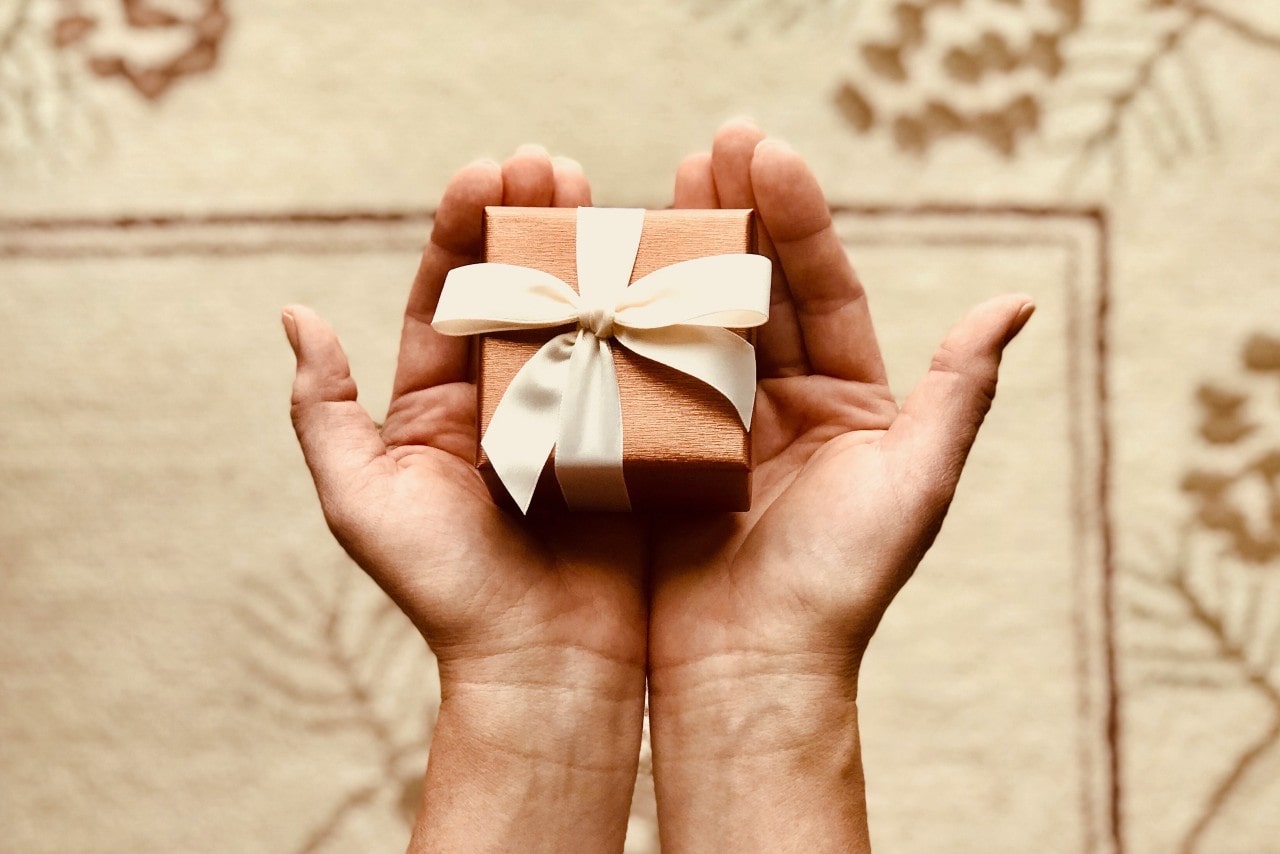 Hands holding a small gift box wrapped in textured paper with a cream ribbon bow, set against a patterned carpet background.