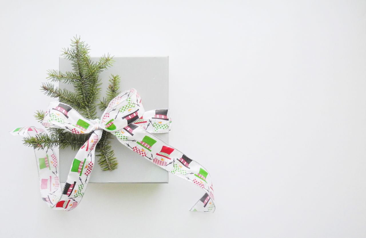 A white gift box with pine sprigs and a festive ribbon featuring colorful patterns lies on a white surface.