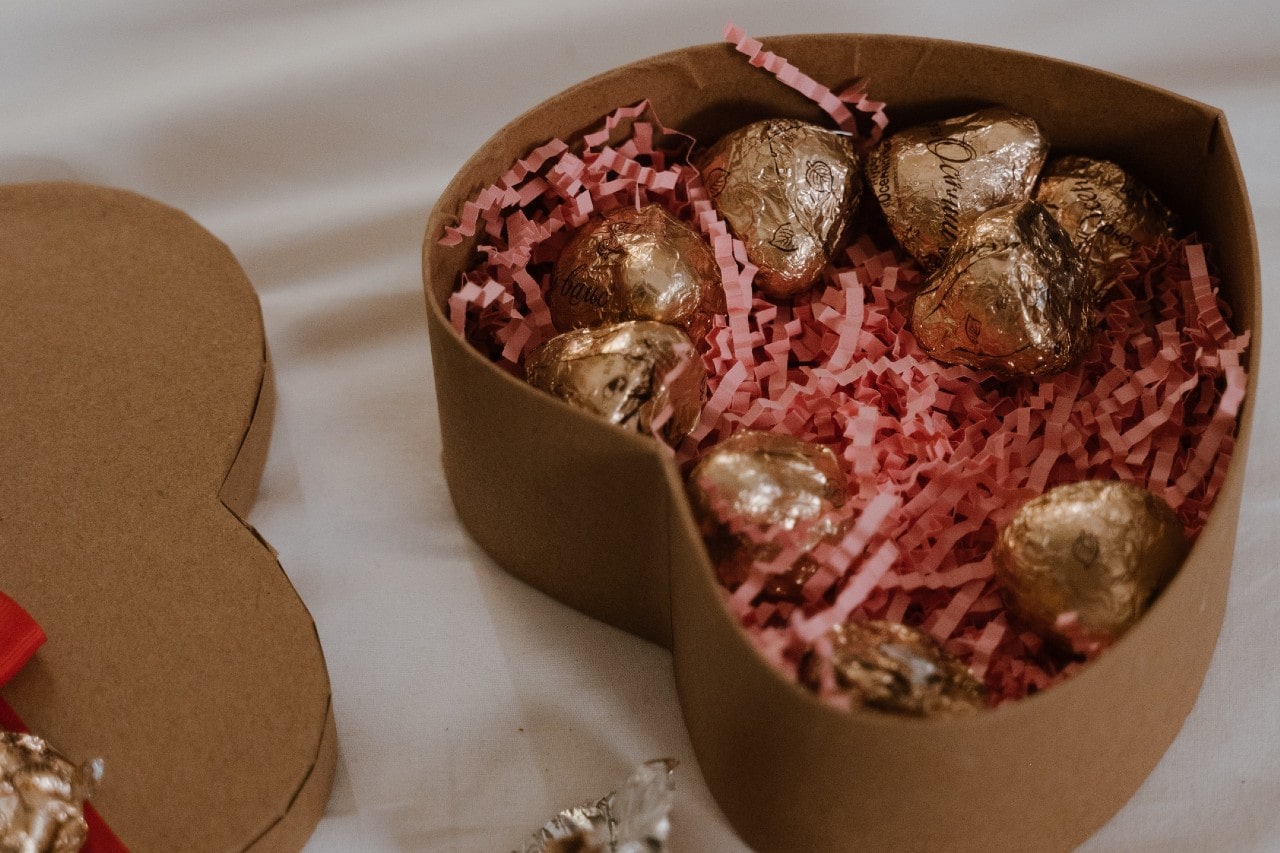 A heart-shaped box filled with pink paper shreds and gold-wrapped chocolates sits open on a white surface.