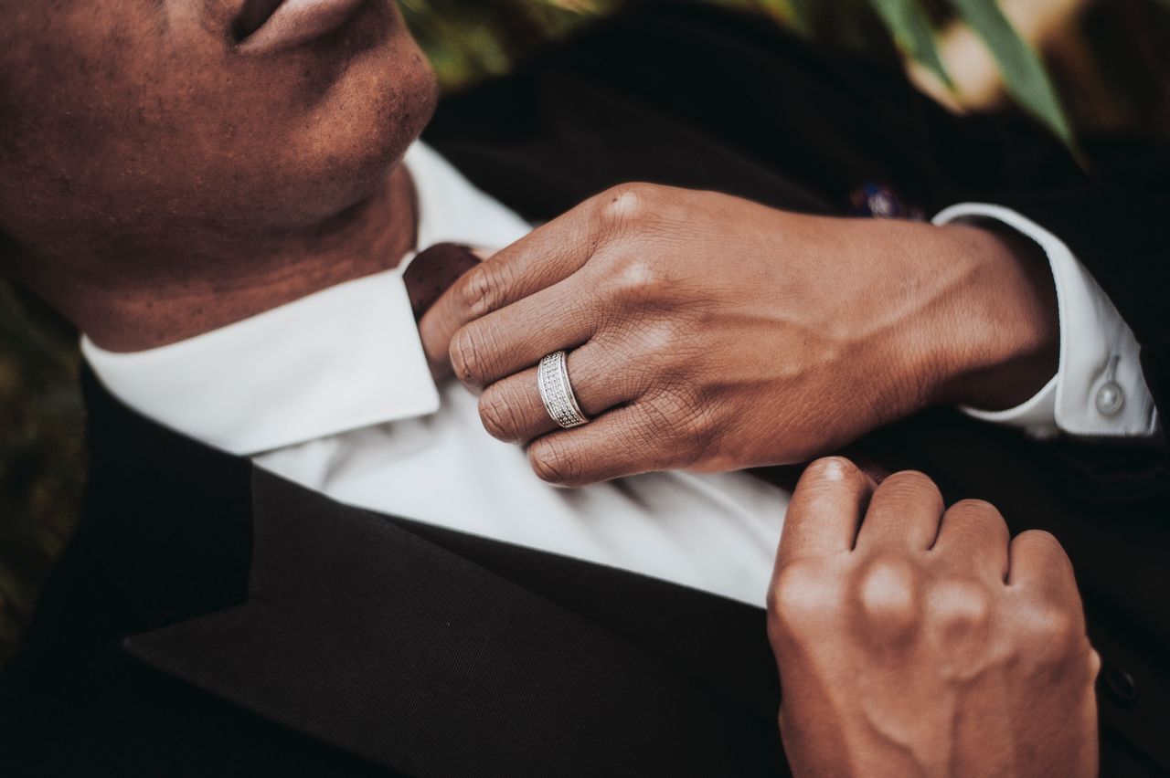 A man in a black suit adjusts his tie, showcasing a wedding band on his finger.