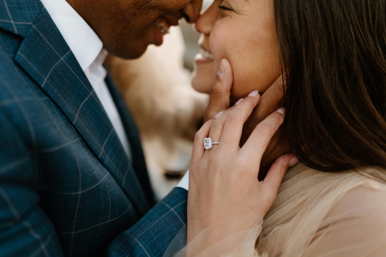 A close-up of a joyful couple leaning towards each other, highlighting the woman's cushion cut engagement ring as she touches her partner's hand.