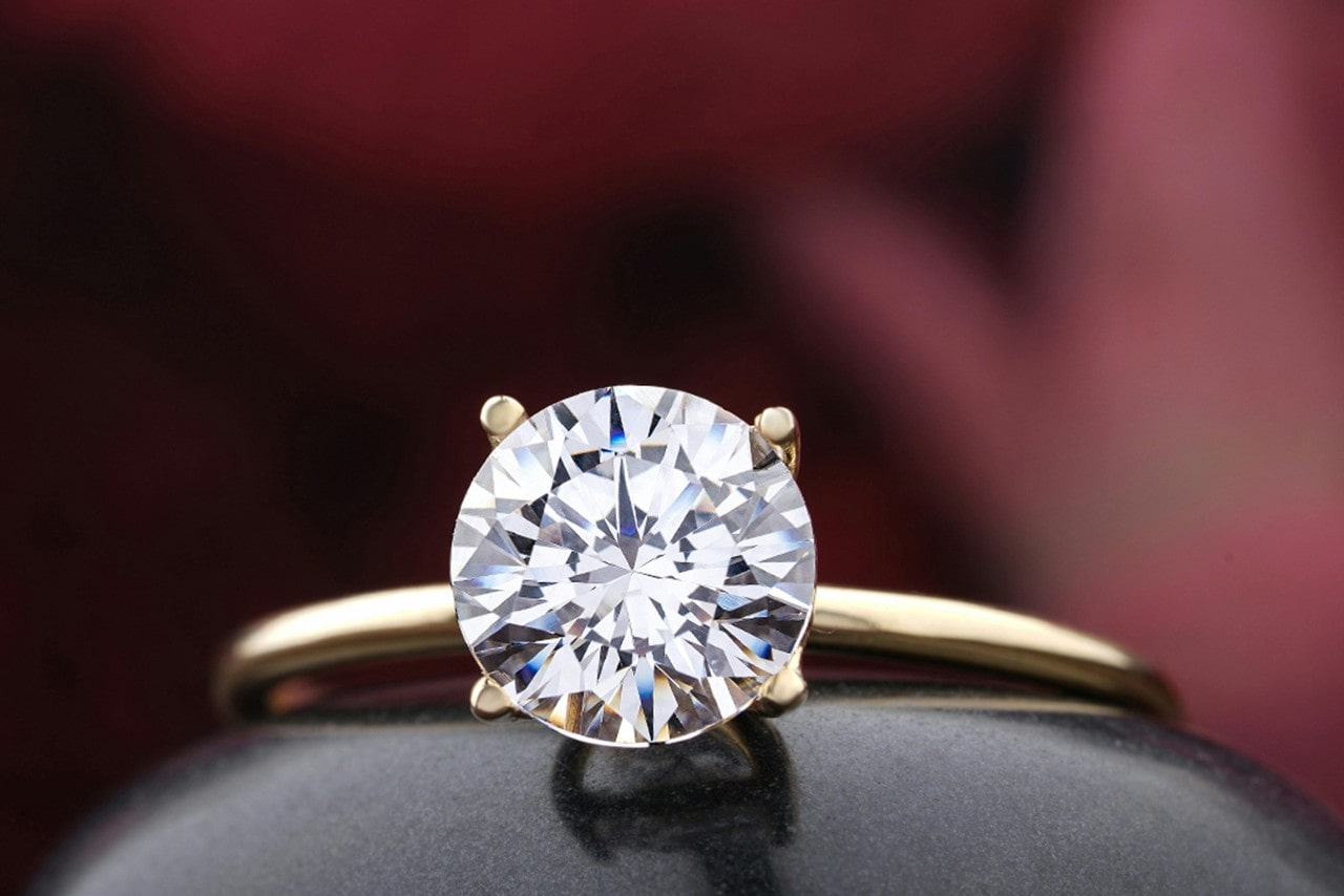 A close-up of a round cut yellow gold thin solitaire engagement ring against a blurry red background.