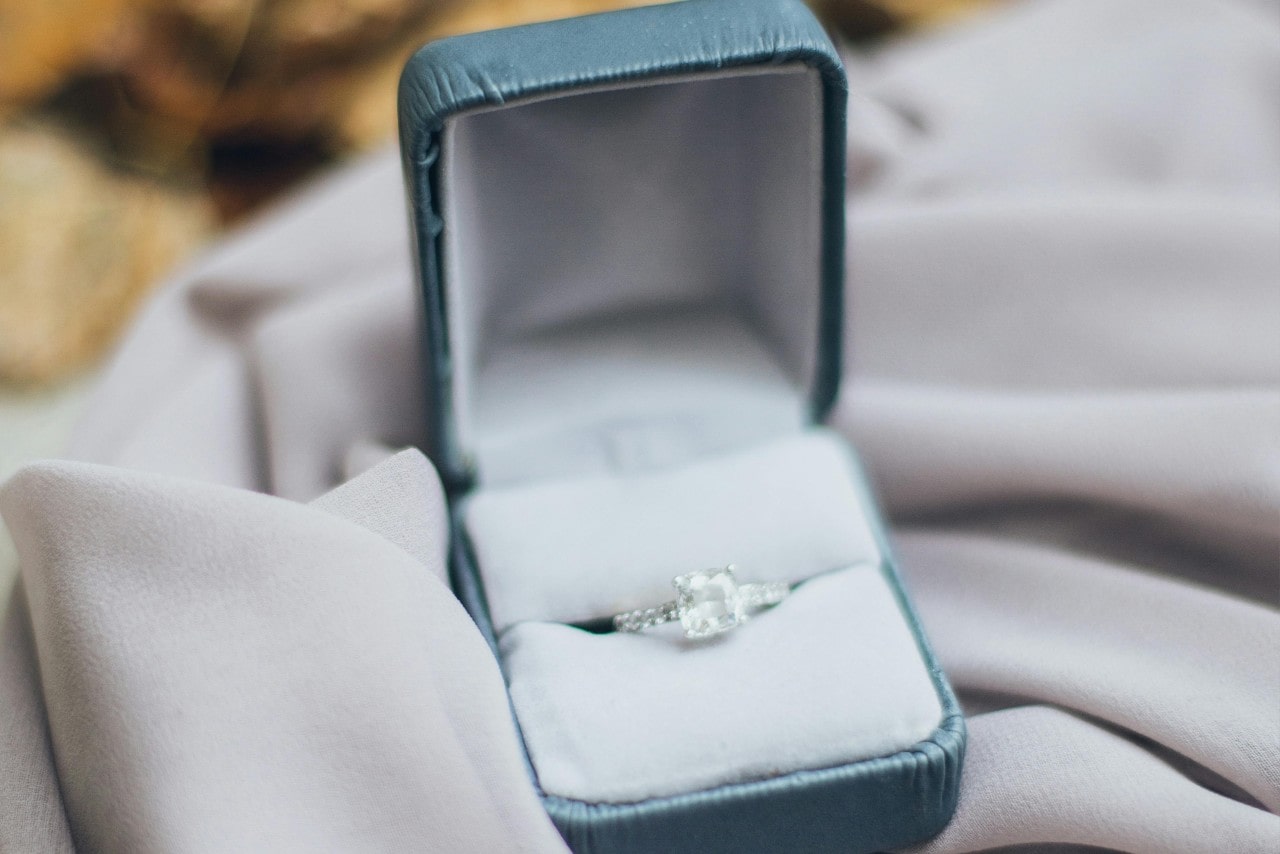 A close-up of a white gold princess cut engagement ring in a green velvet box on a piece of white fabric.