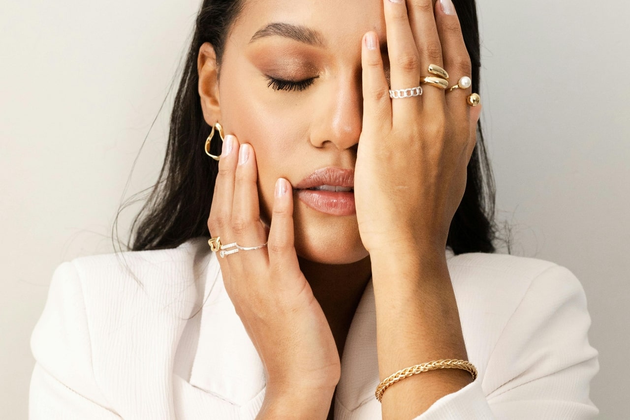 A woman adorned in yellow gold jewelry posing with her hands over her face.