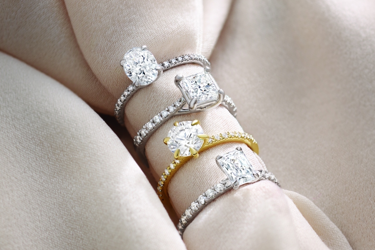 Close-up of a woman's hand with brown nails holding a diamond engagement ring against a blurred background.