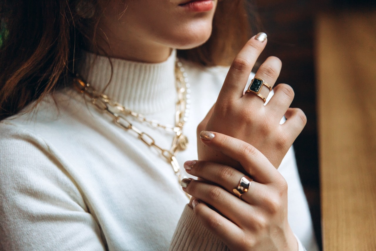 A woman in white turtleneck, adorned with a yellow gold chain necklace and matching fashion rings.
