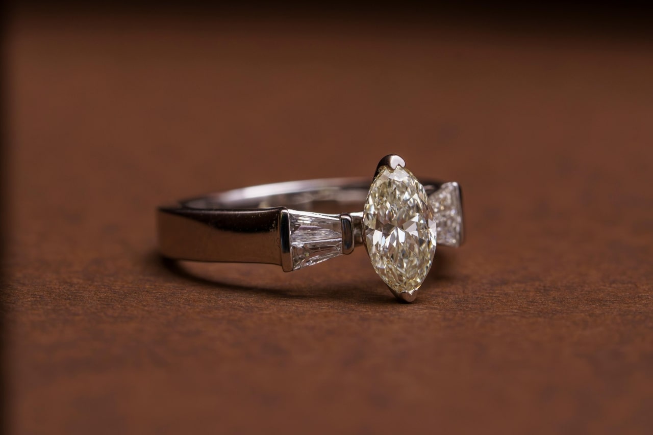 A close-up of a distinctive white gold engagement ring with a marquise cut center diamond on a brown backdrop.