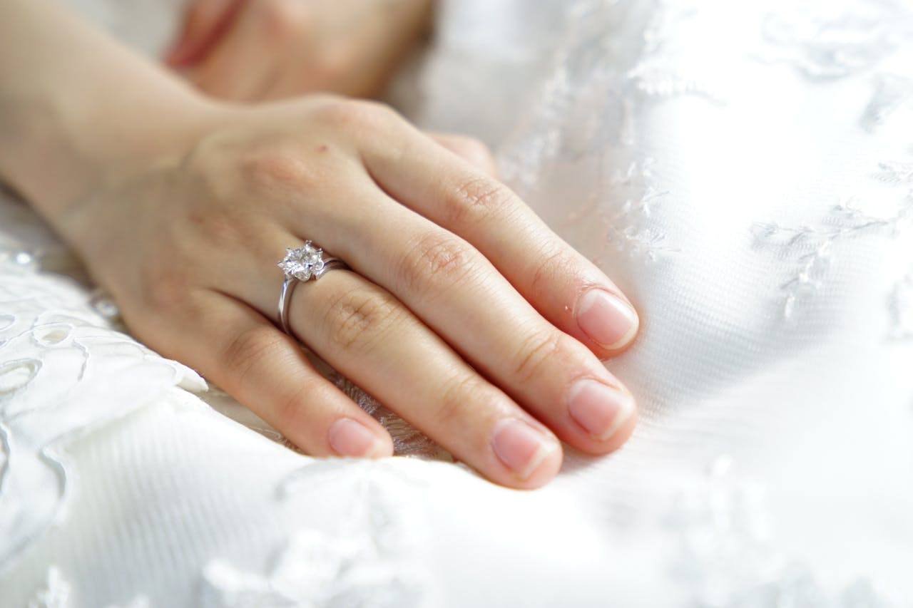 A close-up of a bride’s hand resting on her wedding dress, a simple white gold solitaire engagement ring visible on her finger.