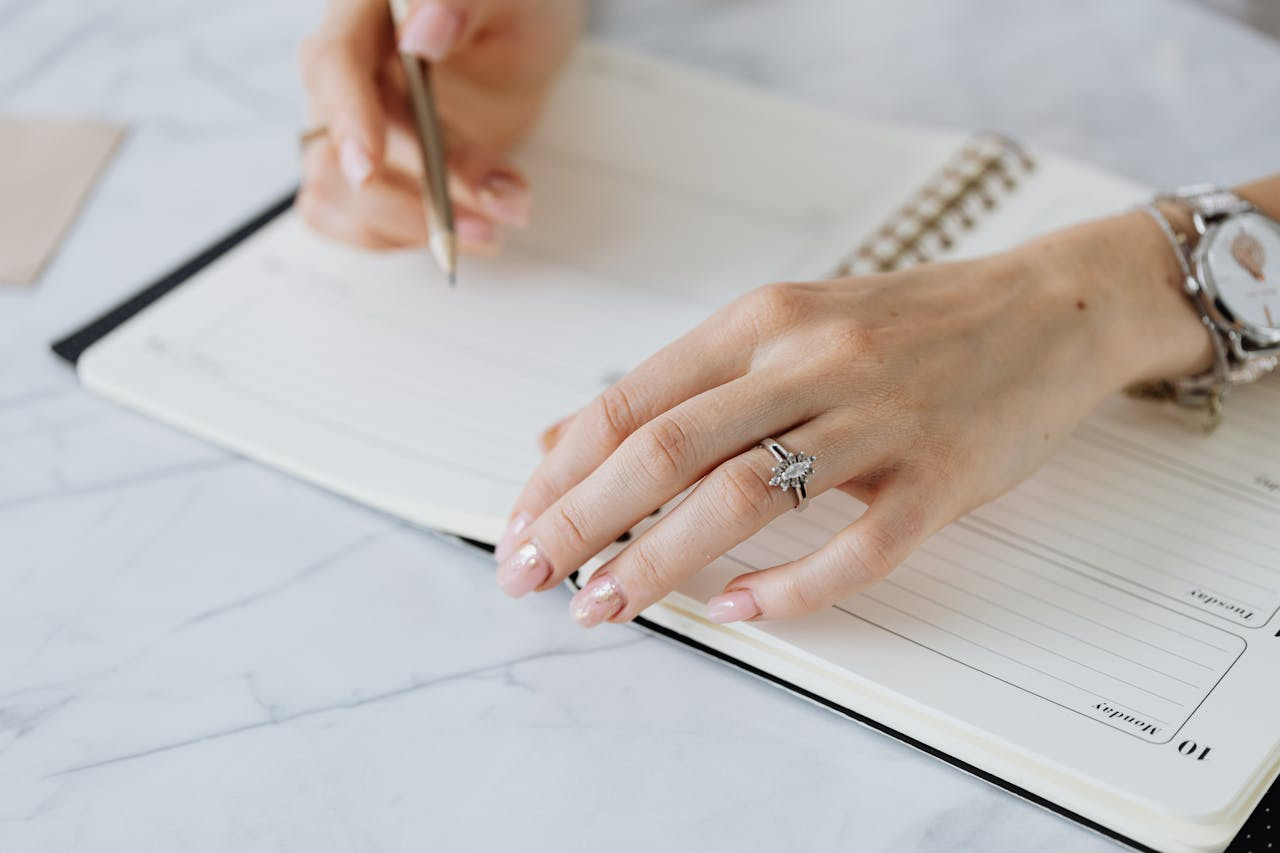 A view of a woman’s hands as she writes in a planner, a distinctive white gold engagement ring visible on her finger.