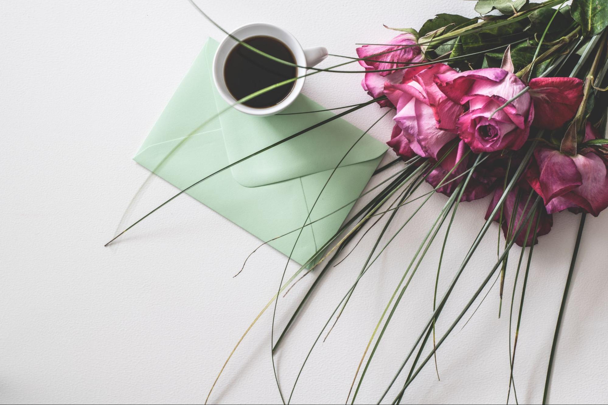 A bouquet of pink roses and long grass beside a green envelope and a cup of black coffee on a white surface.