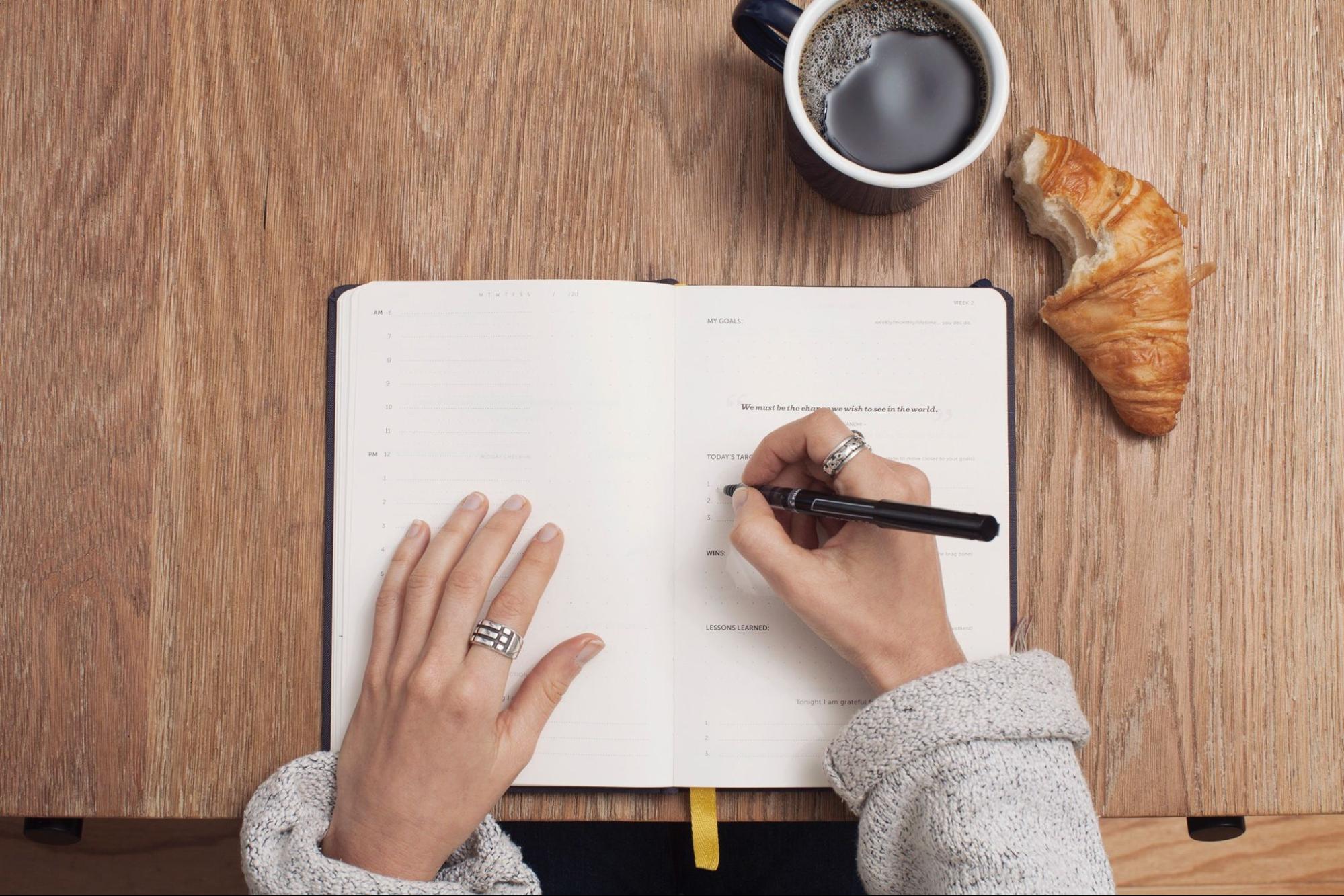 A hand holding a black pen writing something in the open notebook with cup of coffee and a half-bitten croissant lying at the side of a wooden table.