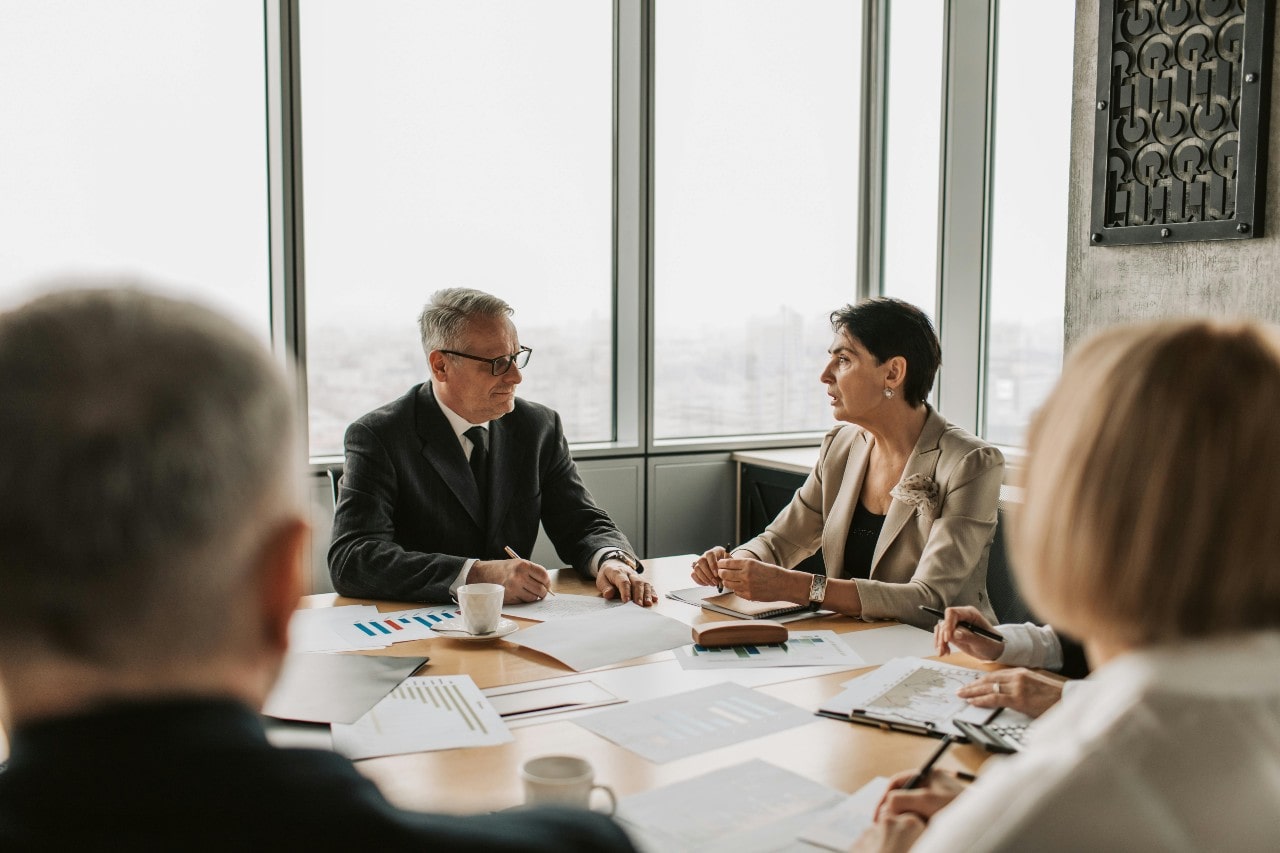 A meeting room where five people sit at a round wooden table on which papers with graphs are scattered, focused on men and women in suits discussing something.