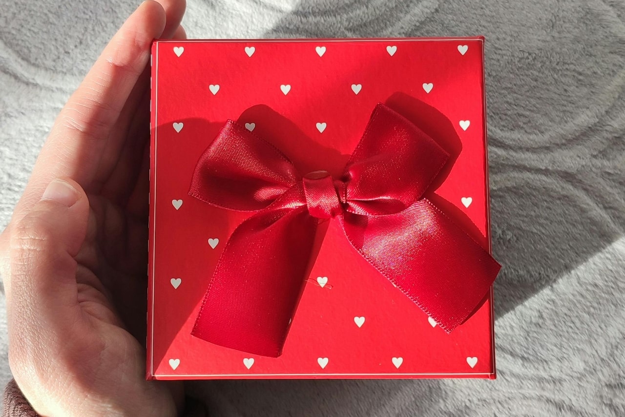 A close-up of a hand holding a square red gift box that has a pattern of little white hearts along with a red silk ribbon at the top.