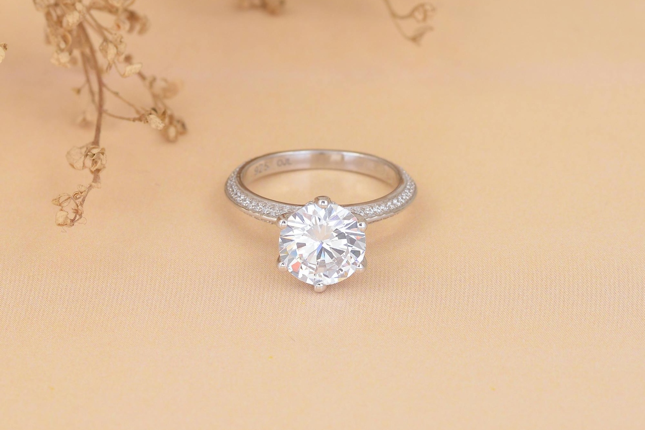 A close-up image of a round brilliant solitaire engagement ring with a pavé diamond band resting on soft beige fabric, surrounded by delicate dried flowers for a romantic and elegant presentation. A close-up image of a round brilliant solitaire engagement ring with a pavé diamond band resting on soft beige fabric, surrounded by delicate dried flowers for a romantic and elegant presentation.