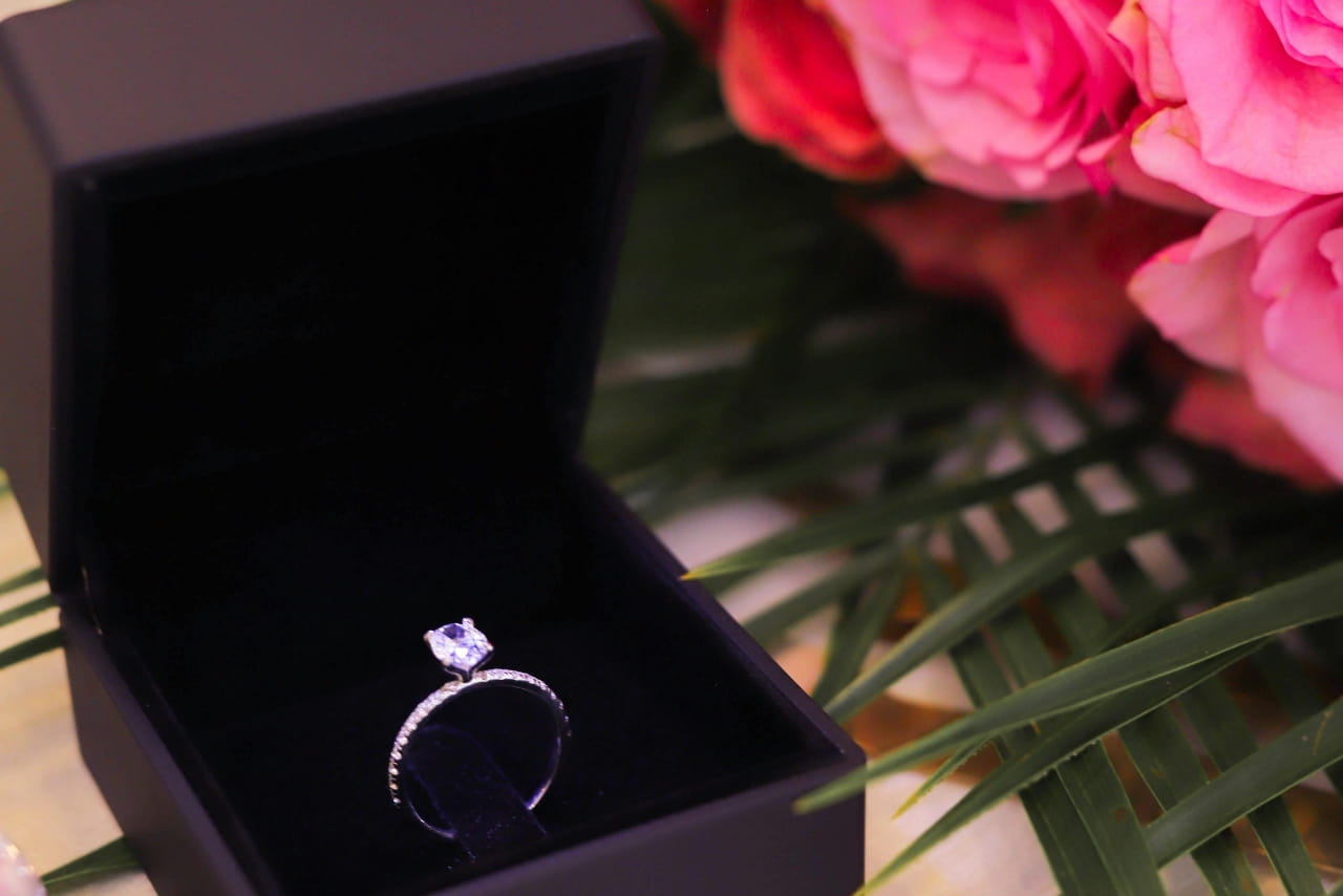 A sparkling engagement ring displayed inside an open black jewelry box, styled with vibrant pink roses and tropical greenery in the background for a luxurious proposal-inspired scene. A sparkling engagement ring displayed inside an open black jewelry box, styled with vibrant pink roses and tropical greenery in the background for a luxurious proposal-inspired scene.