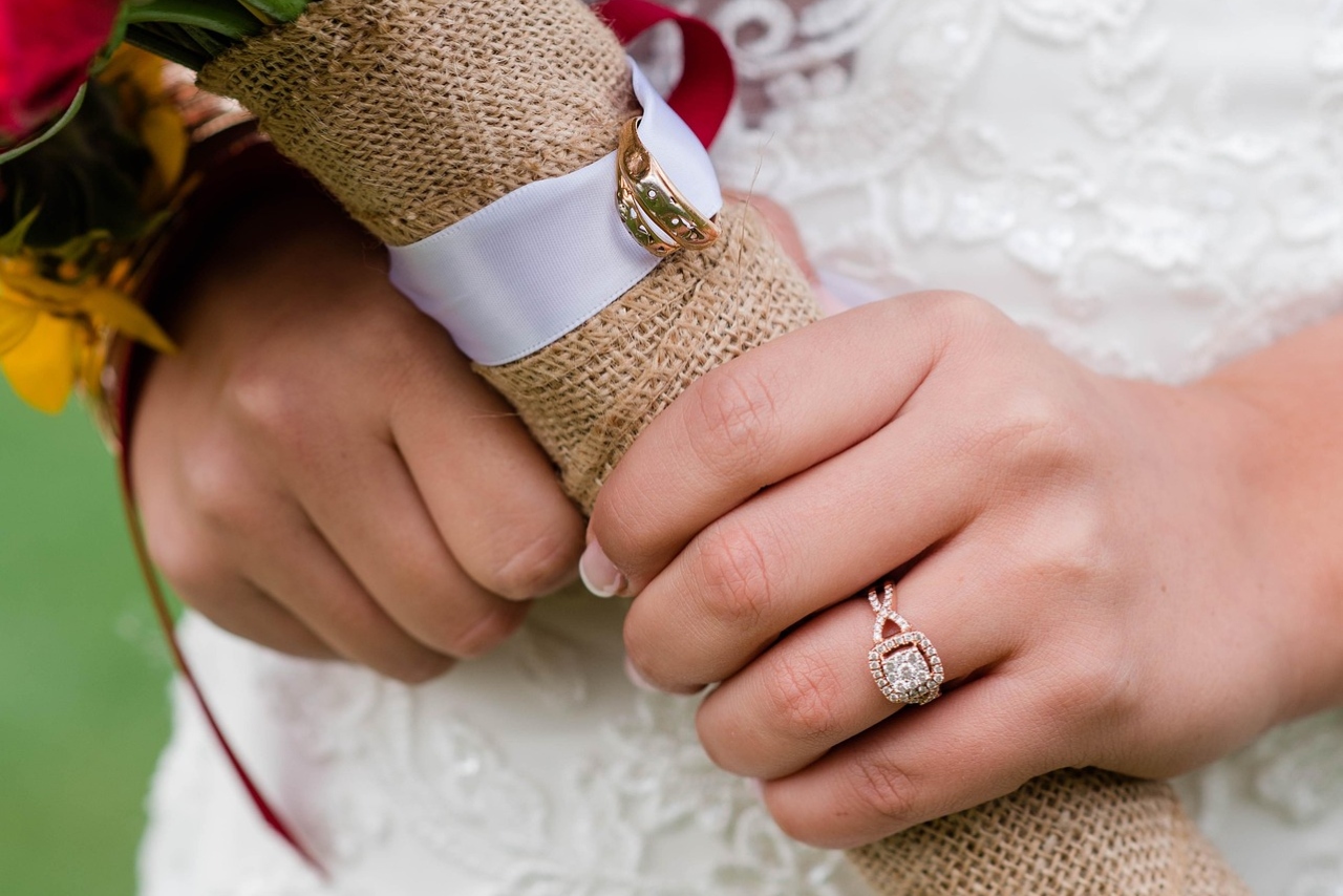 A close-up bridal portrait featuring a bride holding a burlap-wrapped bouquet while wearing a rose gold halo engagement ring and matching wedding band against lace wedding attire. A close-up bridal portrait featuring a bride holding a burlap-wrapped bouquet while wearing a rose gold halo engagement ring and matching wedding band against lace wedding attire.