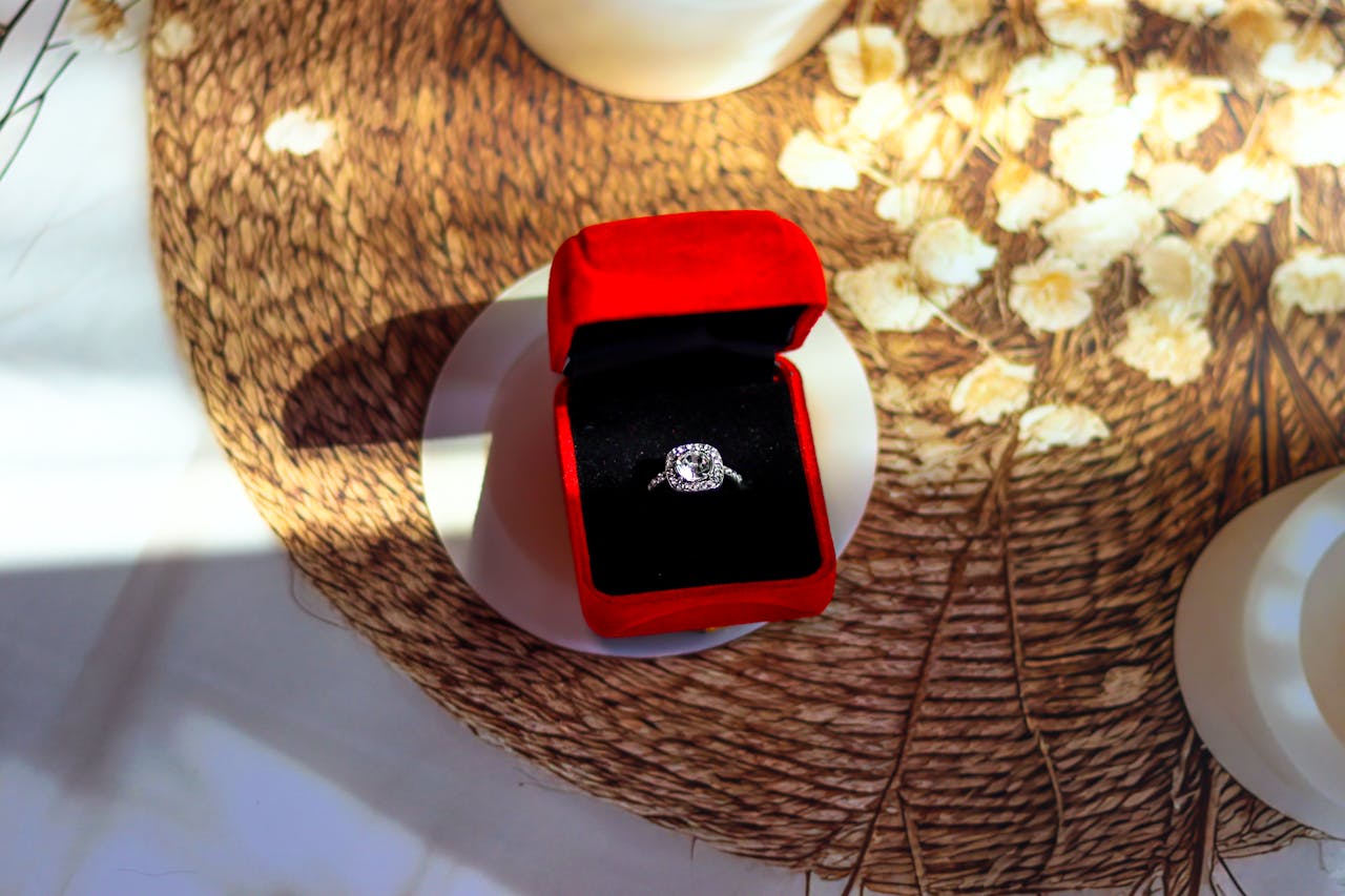 A halo-style diamond engagement ring showcased inside a rich red velvet ring box, placed on a woven textured surface with soft floral accents and dramatic lighting. A halo-style diamond engagement ring showcased inside a rich red velvet ring box, placed on a woven textured surface with soft floral accents and dramatic lighting.