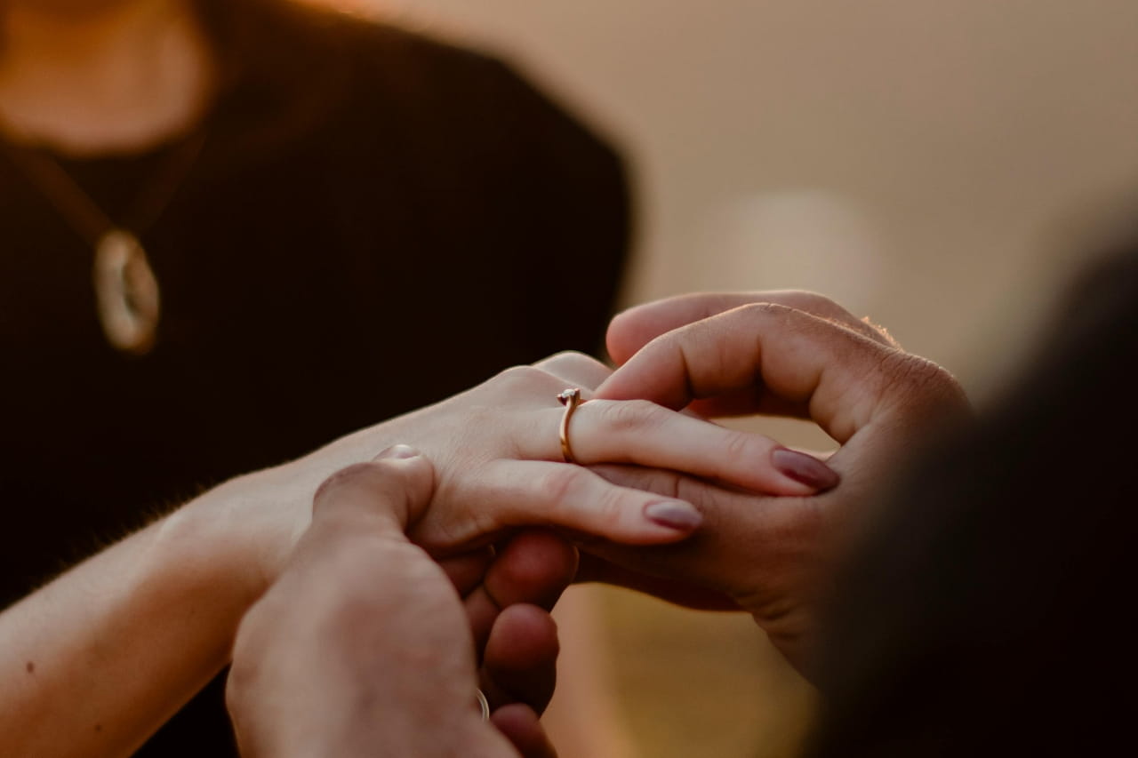 A candid proposal moment showing a person gently placing a delicate engagement ring onto their partner’s finger in warm natural lighting, capturing intimacy and emotion. A candid proposal moment showing a person gently placing a delicate engagement ring onto their partner’s finger in warm natural lighting, capturing intimacy and emotion.