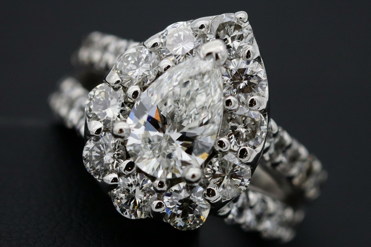 A close-up of an opulent halo engagement ring with a pear cut center stone and a split shank band covered in side diamonds. A close-up of an opulent halo engagement ring with a pear cut center stone and a split shank band covered in side diamonds.