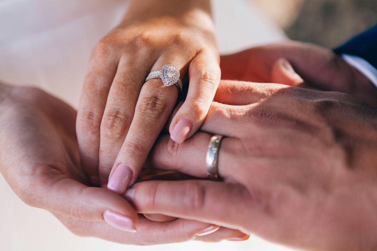 A close-up of a newlywed couple’s hands, with emphasis on the bride’s opulent halo engagement ring. A close-up of a newlywed couple’s hands, with emphasis on the bride’s opulent halo engagement ring.