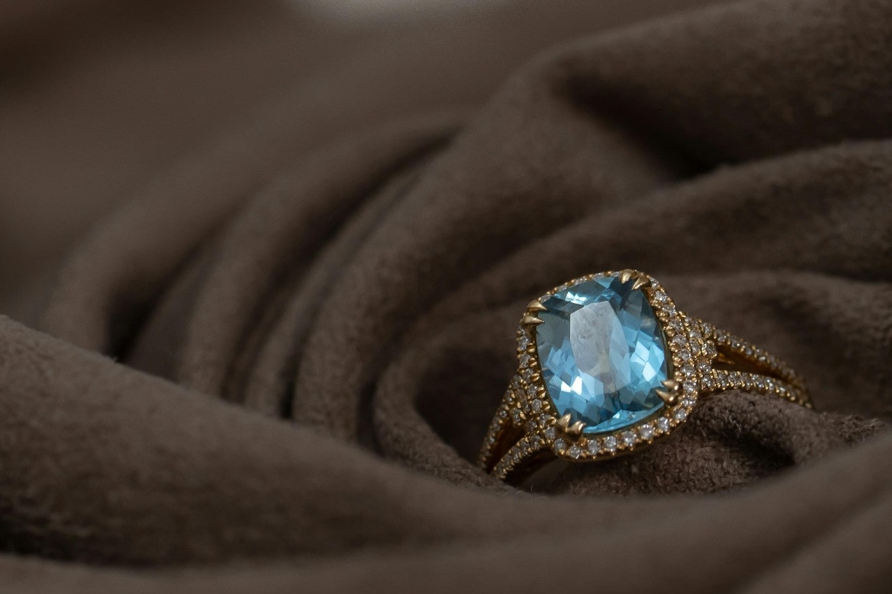 A sparkling blue gemstone ring with a diamond-encrusted gold band rests elegantly on soft, brown fabric. A sparkling blue gemstone ring with a diamond-encrusted gold band rests elegantly on soft, brown fabric.