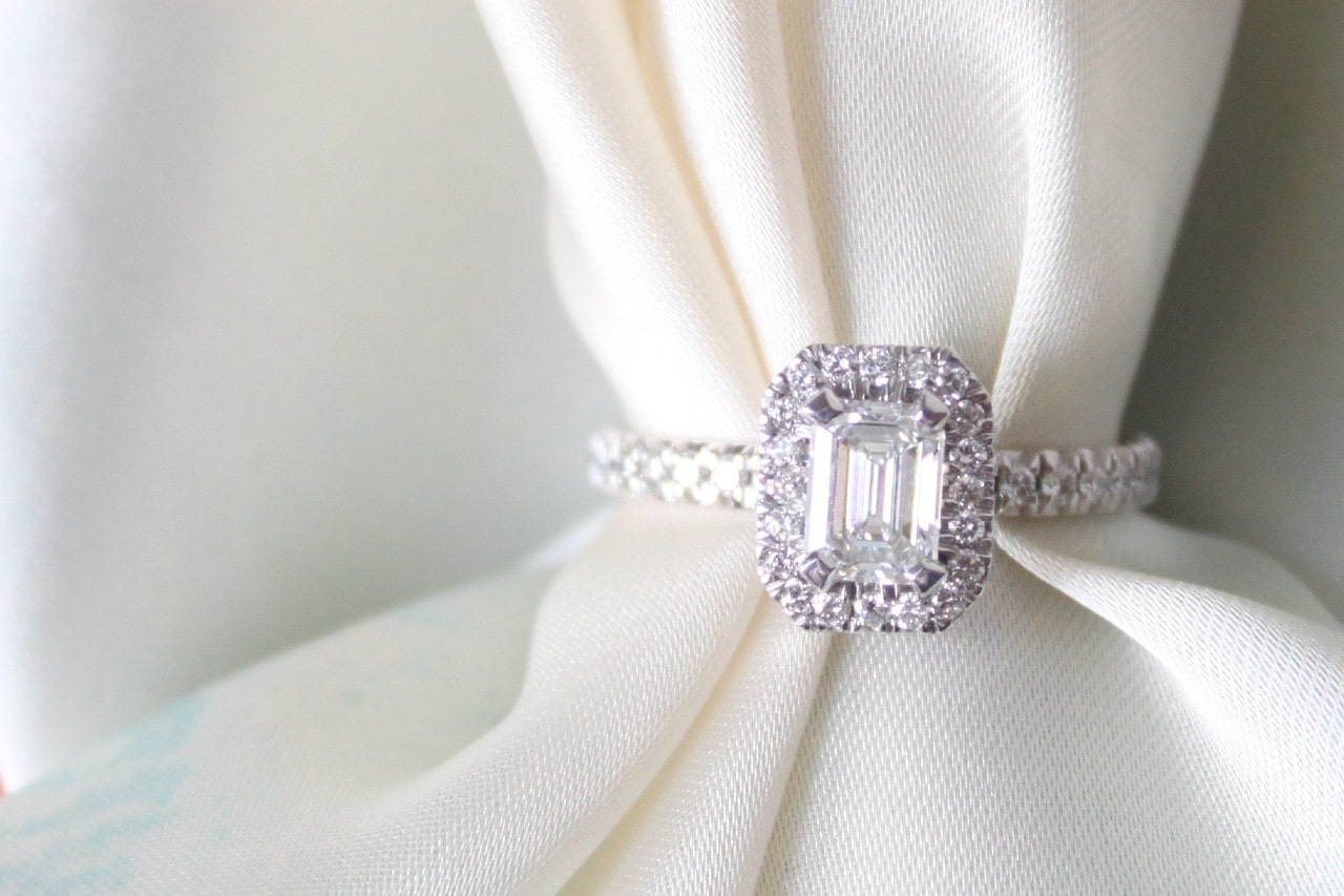 A halo emerald-cut diamond engagement ring on a soft ivory fabric. A halo emerald-cut diamond engagement ring on a soft ivory fabric.