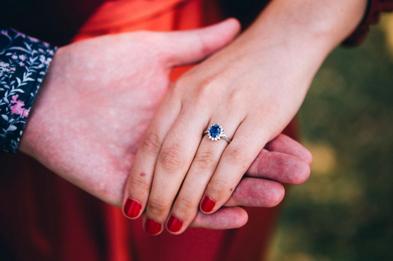 A close up of a woman's hand in an oval-cut halo sapphire engagement ring on the palm of a man's hand in a flower shirt. A close up of a woman's hand in an oval-cut halo sapphire engagement ring on the palm of a man's hand in a flower shirt.