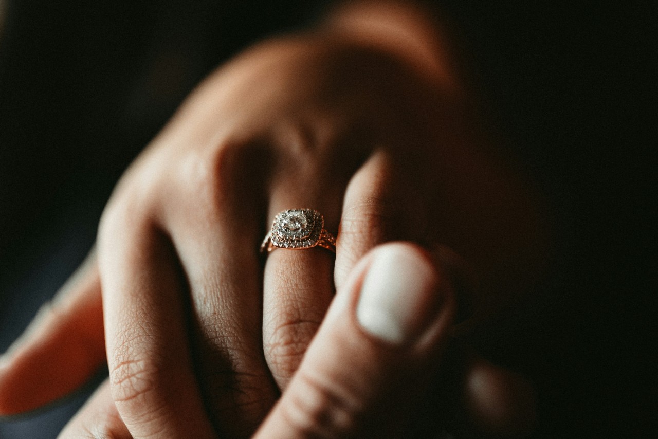 A close-up of a hand wearing a cushion-cut diamond engagement ring, surrounded by a halo of smaller diamonds and held by another hand. A close-up of a hand wearing a cushion-cut diamond engagement ring, surrounded by a halo of smaller diamonds and held by another hand.