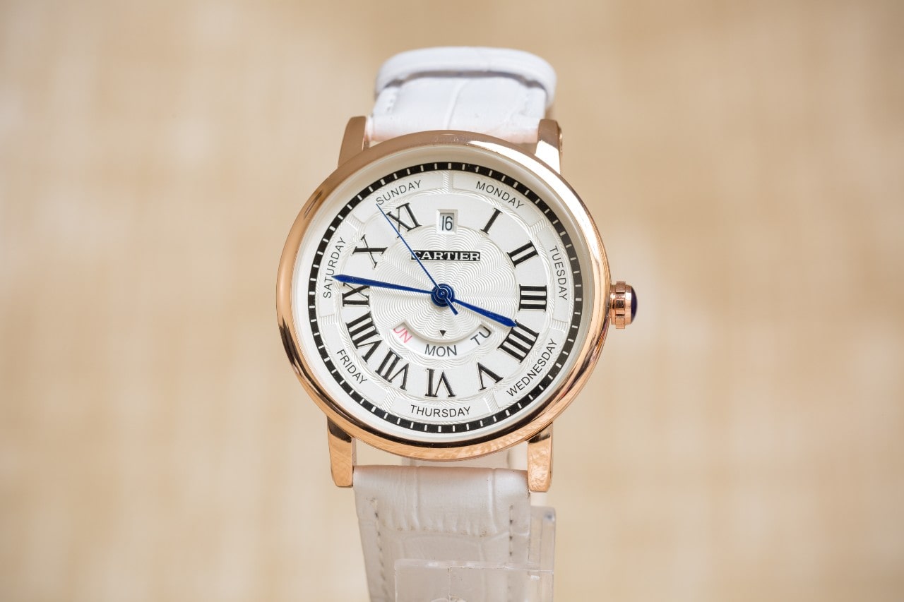 A close-up of a Cartier wristwatch with a white strap, Roman numeral dial, and day-date indicators.