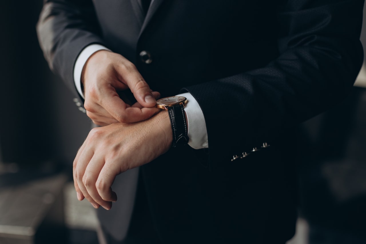 A man in a dark suit adjusts a classic wristwatch with a leather strap on his wrist.