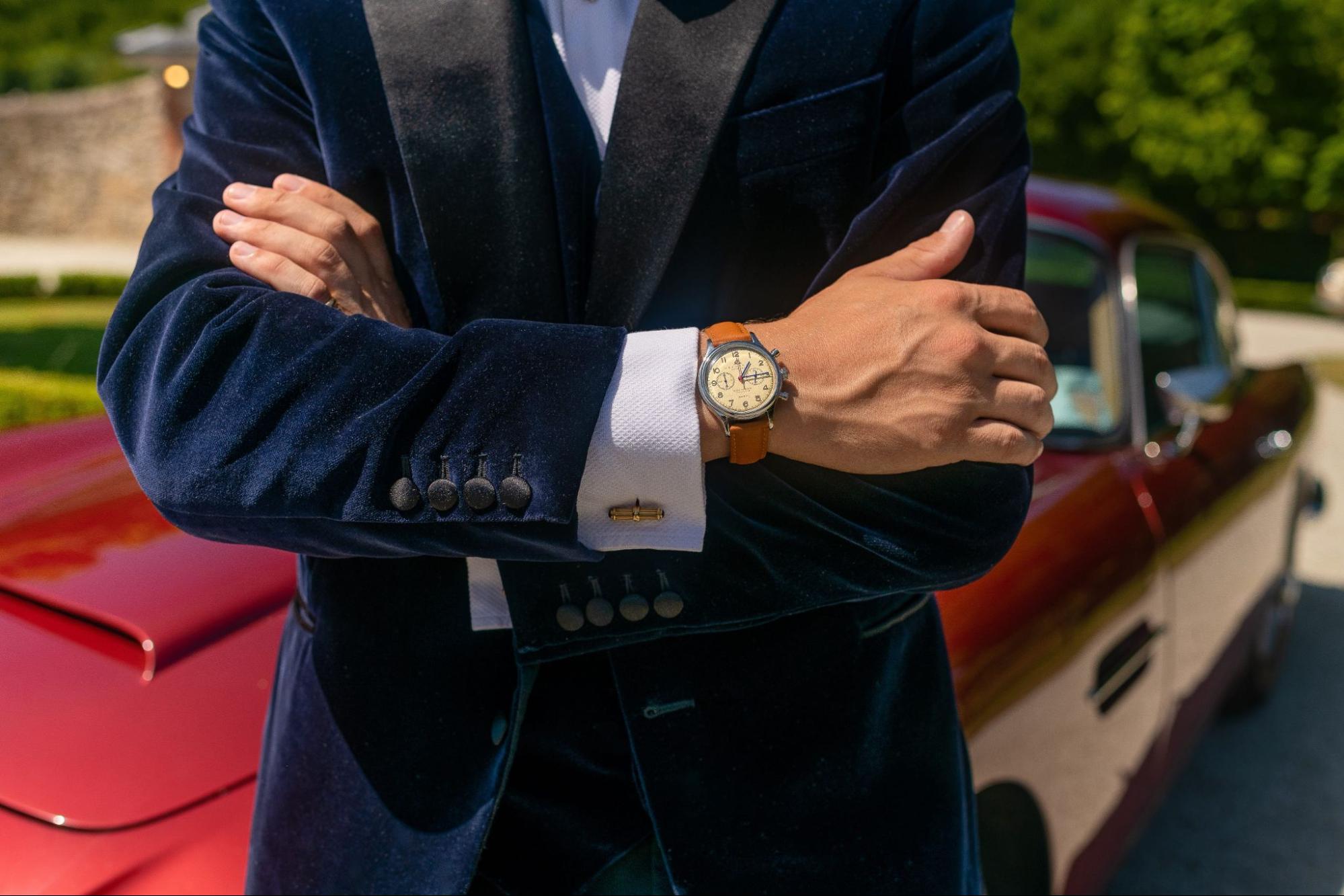 A close up of a man wearing a blue velvet suit with brown leather wristwatch, standing against a red car.