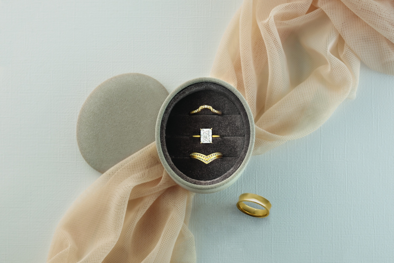 An engagement ring, nesting band, and crowning band displayed in an oval ring box with a matching yellow gold groom’s ring to the side of the decorative display