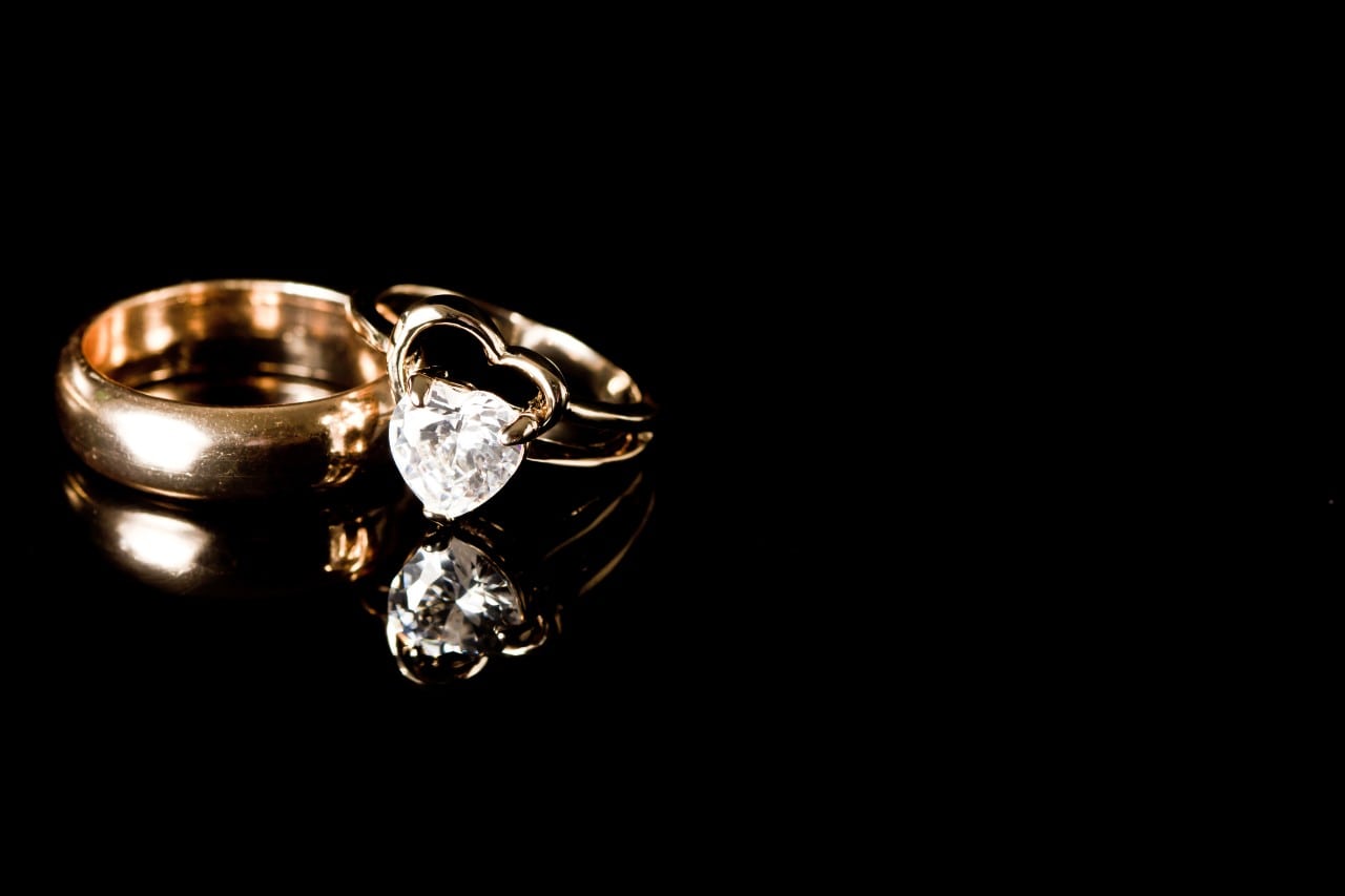 Two gold rings, one with a heart-shaped diamond, rest on a stark black surface. Two gold rings, one with a heart-shaped diamond, rest on a stark black surface.