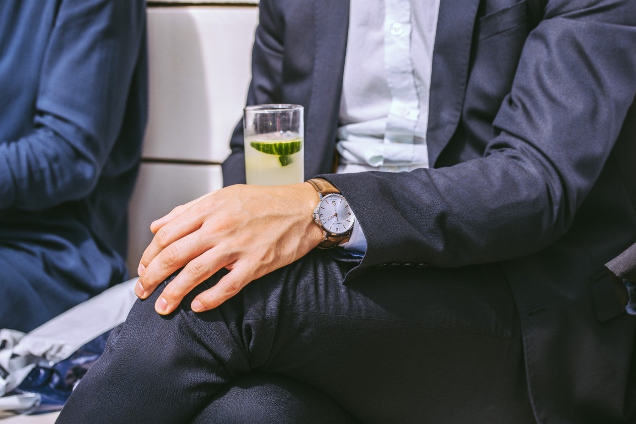 A close-up of a man in a dark blue suit with a glass of lemonade, showcasing a brown leather wristwatch with a white dial.