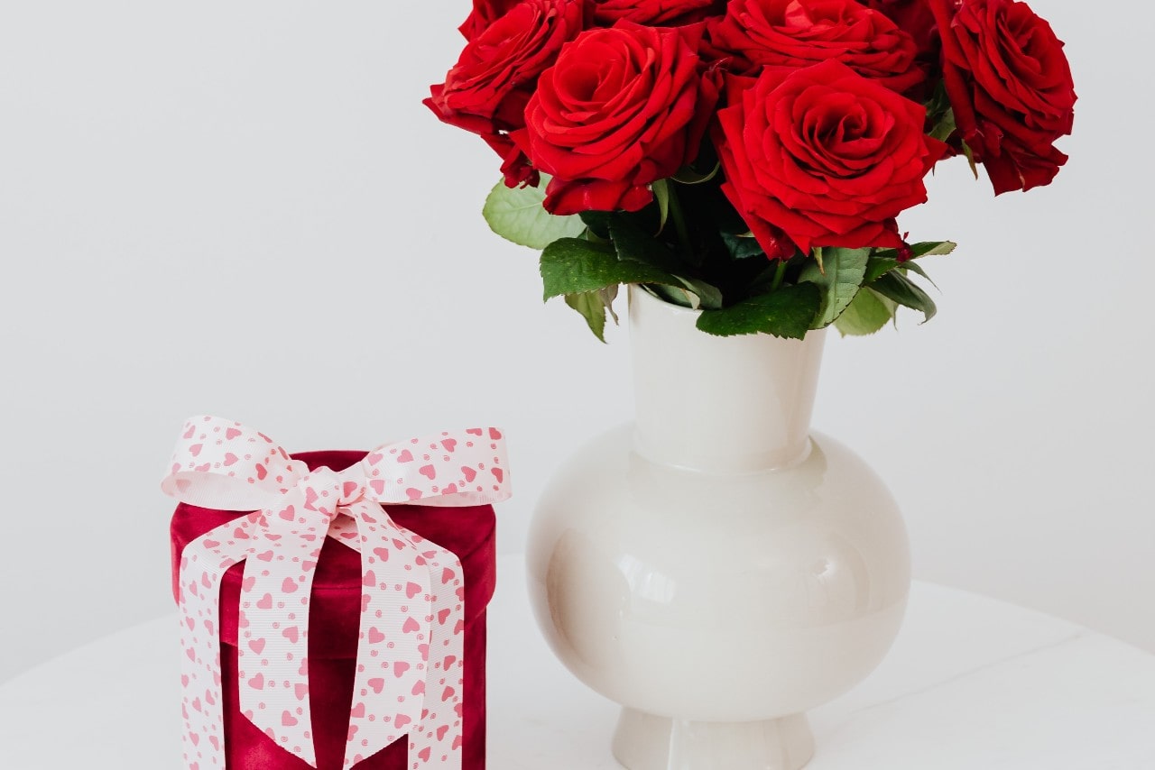 A pink round gift box, wrapped with a white ribbon having pink hearts, next to a white vase with red roses. 